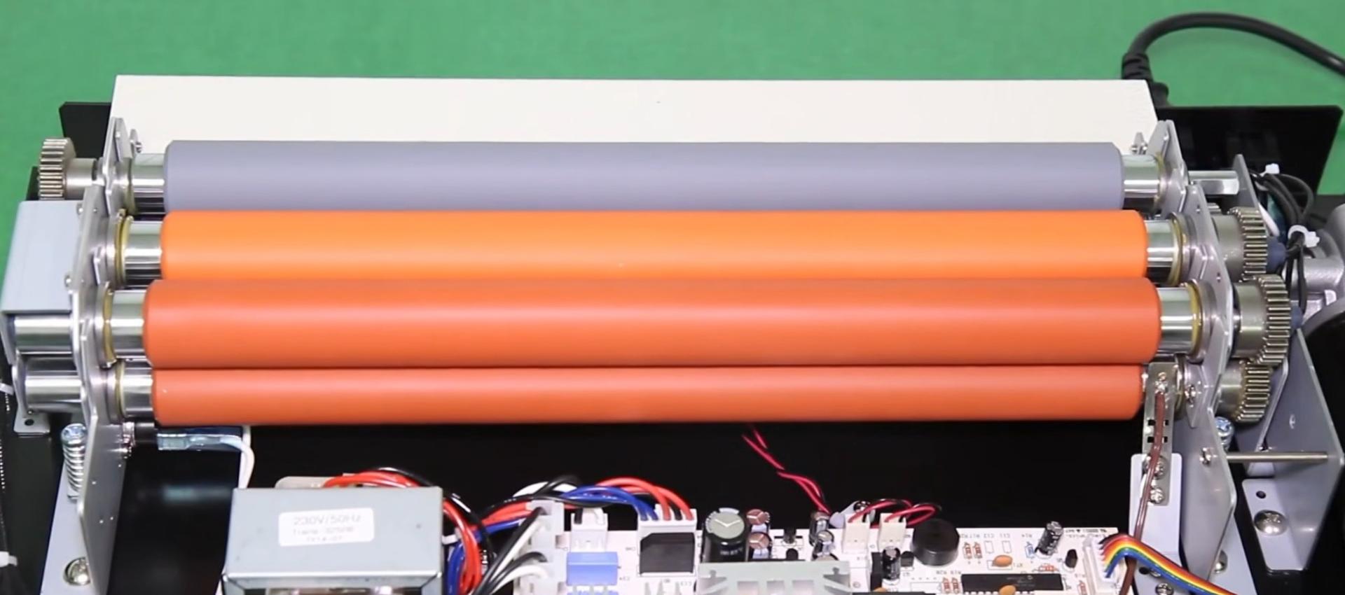 Intérieur d'une plastifieuse à pochettes ouverte : quatre rouleaux superposés montés sur roulements, carte électronique au premier plan